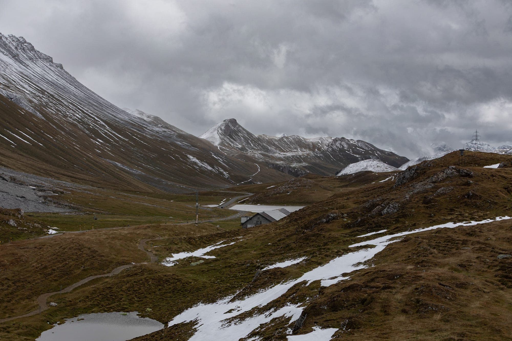 A mountain pass road under the first snow, switchbacking up a bare grey slope beneath low cloud in Graubünden.