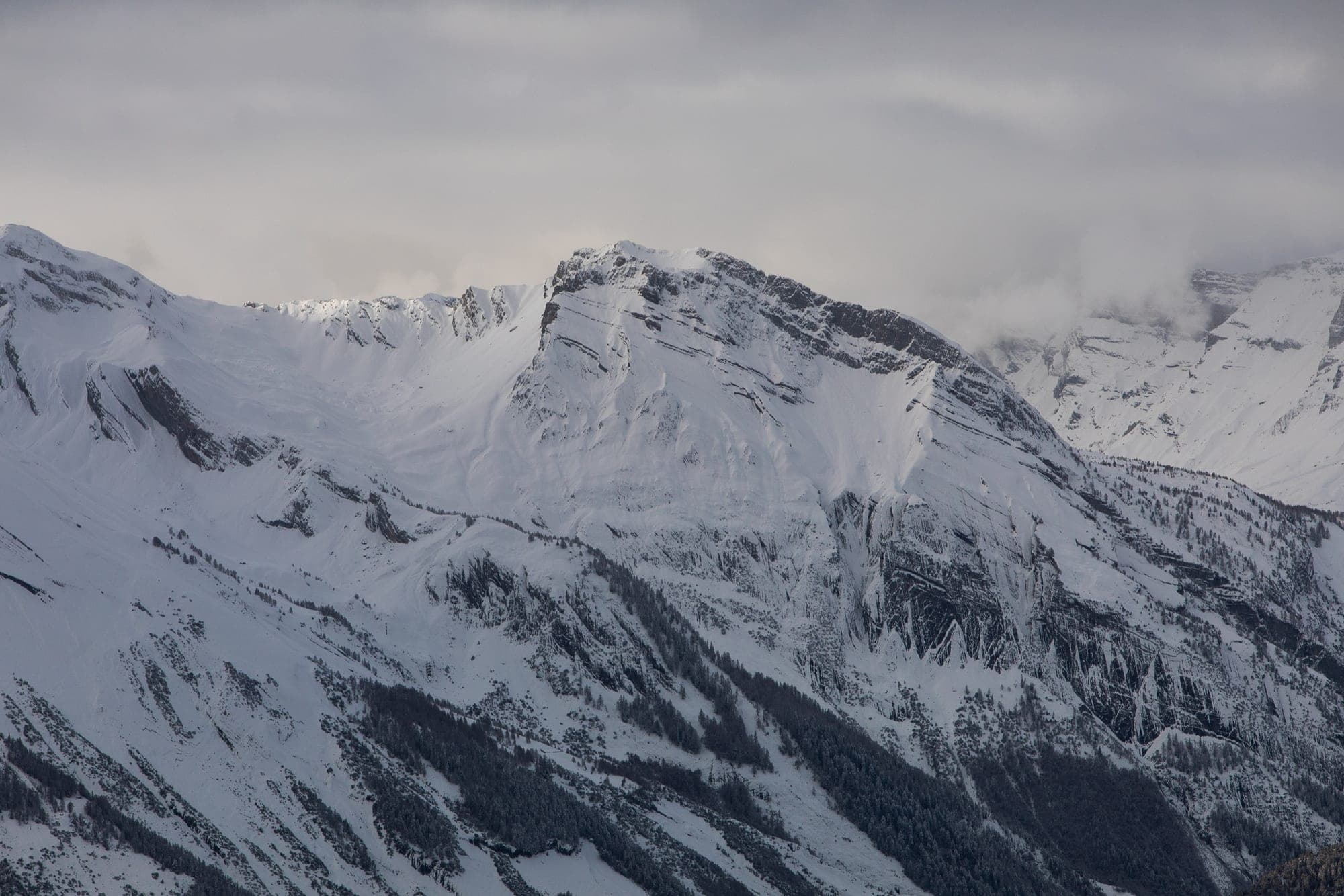 A broad snow massif under solid grey cloud, flat light smoothing its ridges, in Wallis.
