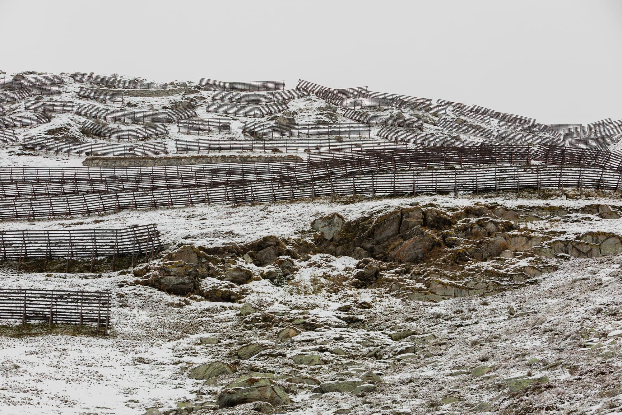 Weathered wooden snow fences crossing a pale snowfield under a flat, overcast sky in Wallis.